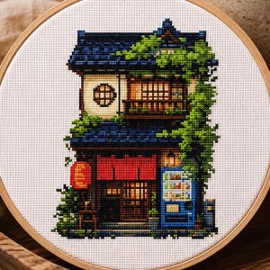 Puede incluir: Un bordado de punto de cruz en un bastidor de madera que representa un edificio japonés de dos plantas con tejado de tejas azul oscuro, enredaderas verdes, un farolillo rojo y una máquina expendedora azul sobre tela blanca cuadriculada.
