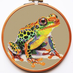 May include: A colorful cross-stitch embroidery of a green, yellow, and orange frog sitting on a beige background. The frog has black spots and a detailed eye.
