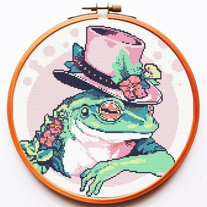 Op de afbeelding: Een groene kikker die een roze en zwarte hoge hoed met bloemen draagt. De kikker zit met gekruiste armen en heeft een roze en groen bloemenontwerp om zijn nek.