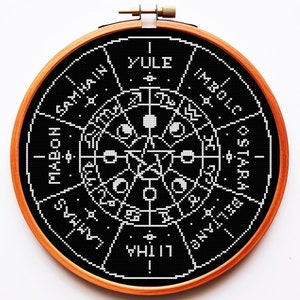 May include: Black and white embroidery hoop with a circular design depicting the Wheel of the Year, a pagan calendar with eight festivals. The festivals are labelled in white text: Yule, Imbolc, Ostara, Beltane, Litha, Lammas, Mabon, and Samhain.