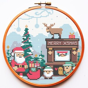 May include: A festive cross-stitch embroidery in a wooden hoop. The design shows a Christmas scene with Santa Claus, a Christmas tree, and a fireplace with the text "MERRY XMAS". A reindeer and gifts are also included in the design.