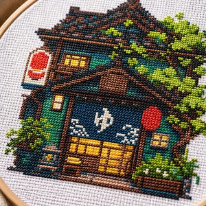 以下が含まれることがあります： 木製の刺繍枠に入ったクロスステッチ刺繍。濃い茶色の屋根瓦、緑の葉、黄色く光る窓がある多層階の建物が描かれています。入り口には青い暖簾がかかっており、白い日本語の文字「ゆ」がプリントされています。