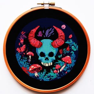 以下が含まれることがあります： 木製のフレームに入ったクロスステッチ刺繍。デザインは、赤い角を持つターコイズのスカル、赤いキノコと暗い青色の背景の葉で囲まれています。アートワークは丸い木製のフレームに収められています。