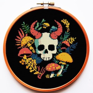 以下が含まれることがあります： オレンジ色の角を持つ骸骨と、カラフルなキノコや葉で囲まれたクロスステッチのデザイン。 黒い背景に配置され、木製の刺繍枠に額装されています。 色のパレットには、ティール、イエロー、レッド、グリーンが含まれています。