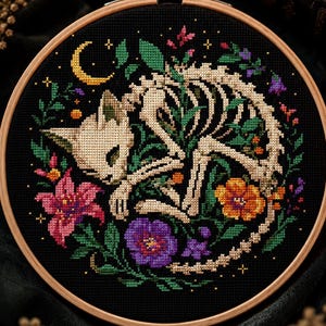 May include: A circular wooden embroidery hoop containing a cross-stitch design on black fabric. The pattern features a beige cat skeleton curled in a sleeping position, surrounded by colorful flowers, green leaves, and a small yellow crescent moon.