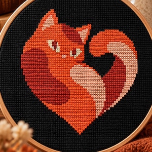 Peut inclure: Un tambour à broder circulaire en bois contenant un motif au point de croix représentant un chat en forme de cœur. Le chat est réalisé dans des tons orange, rouge et crème sur un tissu noir, avec une queue recourbée et de grands yeux.
