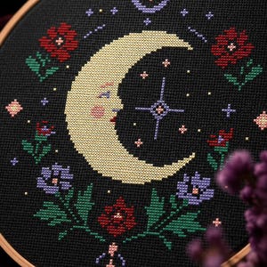 May include: A circular wooden embroidery hoop containing black fabric with a cross-stitch design. The pattern features a large yellow crescent moon with a face, surrounded by red and purple flowers, green leaves, and small stars in various colors.