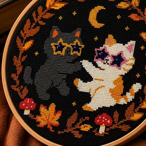 May include: A circular wooden embroidery hoop containing a cross-stitch design on black fabric. The pattern features a grey cat and a white and orange calico cat wearing star-shaped sunglasses, surrounded by autumn leaves, mushrooms, and a crescent moon.