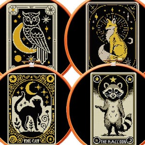 May include: Four circular wooden embroidery hoops containing black fabric with cross-stitch designs. The designs feature an owl with a crescent moon, a fox with a sunburst, a black cat under a moon, and a raccoon with the text THE RACCOON.