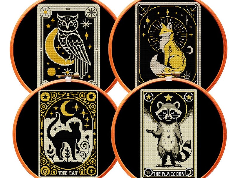 May include: Four circular wooden embroidery hoops containing black fabric with cross-stitch designs. The designs feature an owl with a crescent moon, a fox with a sunburst, a black cat under a moon, and a raccoon with the text THE RACCOON.