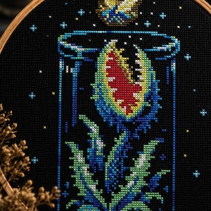 May include: A cross-stitch embroidery in a round wooden hoop featuring a carnivorous plant with a red, yellow, and green mouth inside a blue glass cloche. The design is set against a black fabric background with small white and yellow stars.