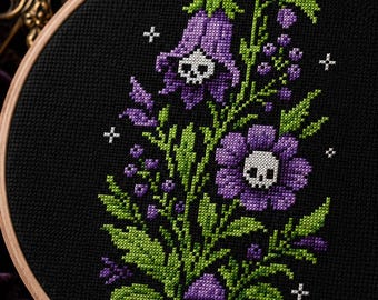 Può includere: Un telaio da ricamo circolare in legno contenente un disegno a punto croce su tessuto nero. Il motivo presenta fiori viola a forma di campana e una margherita viola, ciascuno con un teschio bianco al centro, circondati da foglie verdi e piccole stelle.