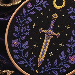 May include: A circular wooden embroidery hoop containing a cross-stitch design on black fabric. The pattern features a central gold and purple sword, a gold crescent moon, small gold stars, and two symmetrical floral vines in shades of purple.