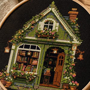 May include: A cross-stitch embroidery in a wooden hoop depicting a small green house with a brown door, a chimney, and windows filled with books. The house is covered in climbing vines and flowers, set against a solid black fabric background.
