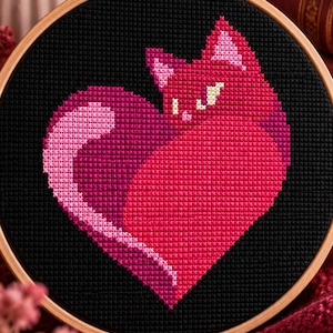 Könnte beinhalten: Ein runder hölzerner Stickrahmen mit einem Kreuzstichmotiv einer herzförmigen Katze. Die Katze ist in verschiedenen Schattierungen von leuchtendem Pink, Magenta und Hellrosa auf einem schwarzen Hintergrund gestickt und hat kleine weiße Augen.