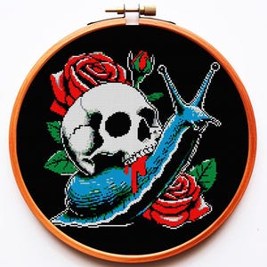 以下が含まれることがあります： 木製のフレームに入ったクロスステッチ刺繍。青いカタツムリに乗った骸骨が描かれ、赤いバラと緑の葉が黒い背景に配置されています。 骸骨には赤い血の一滴があります。ユニークなゴシックアート作品です。