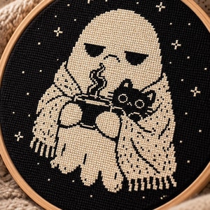 May include: A cross-stitch embroidery piece in a round wooden hoop featuring a cream-colored ghost wrapped in a patterned scarf, holding a steaming mug of coffee, and cuddling a small black cat against a black background with white stars.