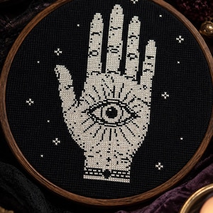 May include: A circular wooden embroidery hoop containing black fabric with a white cross-stitch design of an open palm featuring a central eye and radiating lines, surrounded by small white star-like symbols scattered across the dark background.