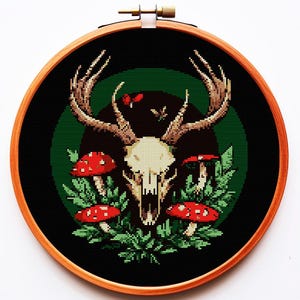 以下が含まれることがあります： 木製のフレームに入ったクロスステッチ刺繍。大きな角を持つ鹿の頭蓋骨、赤いキノコ、緑の葉が黒と緑の背景に描かれています。フレームの直径は約20cmです。
