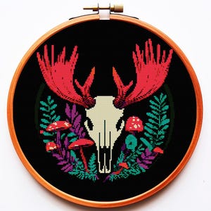以下が含まれることがあります： 黒い背景に、赤い角と緑と紫の葉、赤いキノコが付いた白いヘラジカの頭蓋骨が描かれたクロスステッチ刺繍のフープです。