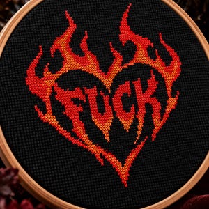 May include: A cross-stitch embroidery piece in a round wooden hoop featuring a heart shape outlined in flames. The design is stitched in red and orange thread on a black fabric background. The word FUCK is centred inside the heart in red.