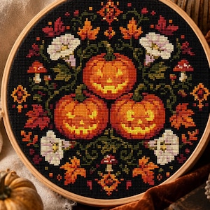 May include: A circular wooden embroidery hoop containing a cross-stitch pattern on black fabric. The design features three orange jack-o'-lanterns surrounded by colorful autumn leaves, small red mushrooms, and white morning glory flowers.