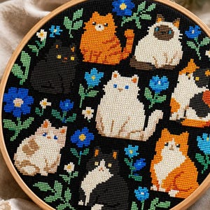 May include: A circular wooden embroidery hoop containing a cross-stitch pattern on black fabric. The design features eight cats of various colors and patterns, including orange, white, black, and calico, interspersed with small blue and white flowers.