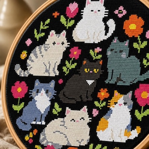 May include: A circular wooden embroidery hoop containing a black fabric cross-stitch pattern featuring six cats in various colors including white, gray, black, and calico, surrounded by colorful embroidered flowers in pink, orange, and red.