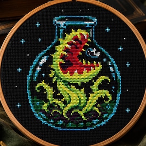 May include: A circular wooden embroidery hoop containing a cross-stitch design on black fabric. The design features a green and red carnivorous plant with tentacles inside a glass jar, surrounded by small white stars against a dark background.