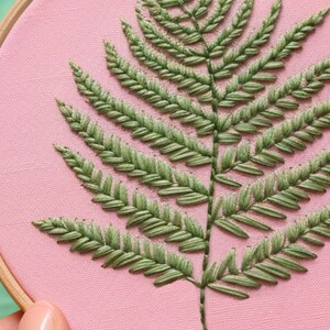 K&ouml;nnte beinhalten: Ein gr&uuml;ner Farn, der auf einem rosa Stoff gestickt ist, der &uuml;ber einen h&ouml;lzernen Stickrahmen gespannt ist.