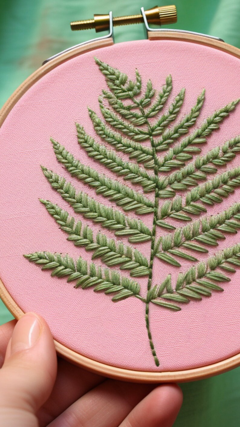 K&ouml;nnte beinhalten: Ein gr&uuml;ner Farn, der auf einem rosa Stoff gestickt ist, der &uuml;ber einen h&ouml;lzernen Stickrahmen gespannt ist.