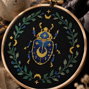 May include: A circular wooden embroidery hoop containing a cross-stitch design on black fabric. The design features a central blue and yellow beetle surrounded by a wreath of green leaves, small blue flowers, and yellow crescent moon and cross symbols.