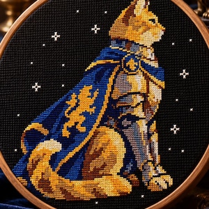 May include: A circular wooden embroidery hoop containing a cross-stitch design of a cat wearing silver armor and a blue cape with a gold lion emblem. The cat is set against a black background featuring small white stitched stars.