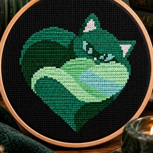 Puede incluir: Un proyecto de punto de cruz en un bastidor de madera circular, que muestra un gato estilizado en forma de corazón. El gato está bordado en varios tonos de verde y azul claro sobre una tela negra, con pequeños detalles en color rosa.