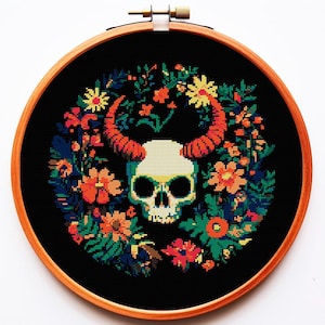 以下が含まれることがあります： 木製のフレームに入ったクロスステッチ刺繍。オレンジ色の角を持つ骸骨が、黒い背景にカラフルな花と葉で囲まれています。フレームは丸く、明るい茶色の木材でできています。