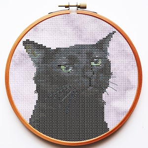 May include: A cross-stitch embroidery of a black cat with green eyes, set within a circular wooden hoop. The cat is depicted against a light purple, mottled fabric background. The embroidery features detailed stitching to define the cat's face.