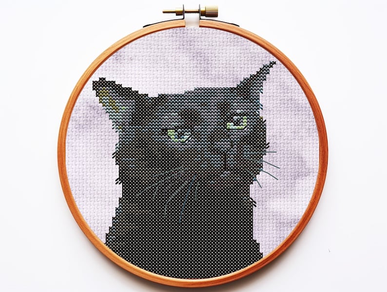 May include: A cross-stitch embroidery of a black cat with green eyes, set within a circular wooden hoop. The cat is depicted against a light purple, mottled fabric background. The embroidery features detailed stitching to define the cat's face.