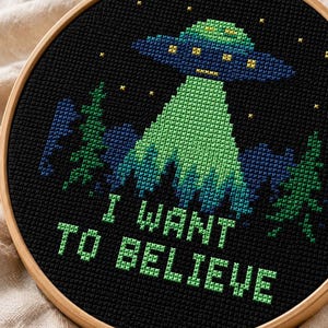 Könnte beinhalten: Ein runder Stickrahmen aus Holz mit einem Kreuzstichmotiv auf schwarzem Stoff. Das Design zeigt ein grünes und blaues UFO über dunkelblauen und grünen Tannenbäumen, ergänzt durch gelbe Sterne und den grünen Schriftzug I WANT TO BELIEVE.