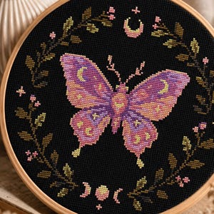 Puede incluir: Un bordado de punto de cruz en un bastidor de madera redondo con una mariposa central de color morado y naranja con motivos lunares. La mariposa está rodeada por una corona circular de hojas verde oscuro y pequeñas flores de color rosa.