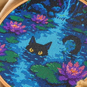 May include: A circular embroidery hoop containing a cross-stitch pattern of a black cat submerged in a blue pond. The scene features vibrant pink water lilies, green lily pads, and a dark blue water surface with a visible cat head and tail.