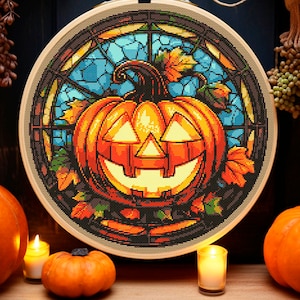 May include: A cross-stitch embroidery of a jack-o'-lantern pumpkin in a stained glass window design. The pumpkin is orange with a bright yellow face and is surrounded by fall leaves.