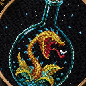 May include: A circular wooden embroidery hoop containing a cross-stitch design on black fabric. The design features a yellow and orange carnivorous plant with a red interior inside a glass bottle, surrounded by small white stars on a dark background.