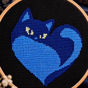 Könnte beinhalten: Ein runder Stickrahmen aus Holz mit einem Kreuzstichmotiv einer Katze, die sich zu einer Herzform zusammengerollt hat. Die Katze ist in Blautönen auf schwarzem Stoff gestickt und hat leuchtend gelbe Augen sowie einen langen, geschwungenen Schwanz.