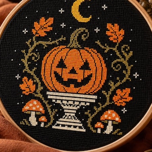 May include: A circular wooden embroidery hoop containing a cross-stitch design on black fabric. The pattern features an orange jack-o'-lantern on a pedestal, surrounded by green vines, orange oak leaves, small mushrooms, and a yellow crescent moon.