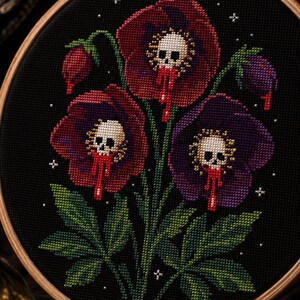 May include: A cross-stitch embroidery piece in a round wooden hoop featuring three dark red and purple flowers on a black background. Each flower center contains a small white skull with red drips, surrounded by green leaves and tiny white stars.