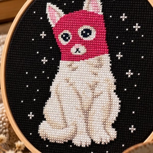 May include: A circular wooden embroidery hoop containing a cross-stitch design of a cat against a black background. The cat has a bright pink head and ears, with a white body featuring light brown shading and small white cross-stitched stars.