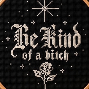 May include: A circular wooden embroidery hoop containing black fabric with white cross-stitch embroidery. The design features a large star at the top, the text "Be Kind of a bitch" in a gothic font, and a single rose at the bottom with small stars.