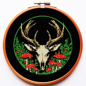 以下が含まれることがあります： 黒と緑の刺繍枠に、白い鹿の頭蓋骨が赤いキノコと緑の葉に囲まれている。