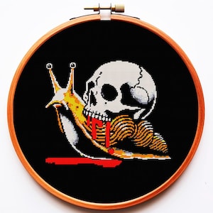 以下が含まれることがあります： 木製のフレームに入ったクロスステッチアート。黒い背景に、カタツムリの体を持つ骸骨が描かれています。カタツムリは黄色と白で、赤い跡があります。骸骨は白で、黒いディテールがあります。