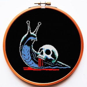 以下が含まれることがあります： 木製のフレームに入ったクロスステッチアート。デザインは、黒い背景に、頭蓋骨の頭と口から赤い血が滴る青いカタツムリが特徴です。カタツムリは青と白のピクセルで詳細に描かれています。